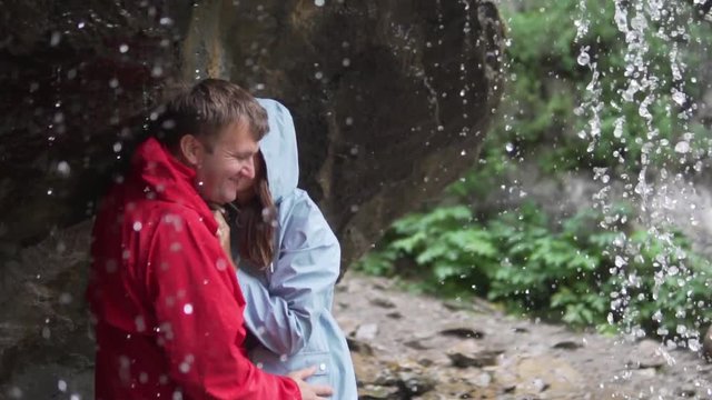 Family day. Lovers kiss under a waterfall. A couple on a hike stands under a stream of a waterfall in non-wet jackets, they gently kiss and laugh