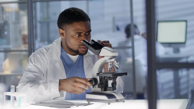 Waist-up shot of middle-aged African American lab technician in medical uniform sitting at work in clinic and studying tissue sample under microscope, looking in repeatedly and filling analysis sheet