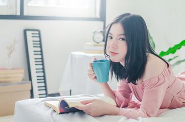 Time for myself. Comfort and relaxation. Pretty young asian woman drinking tea or coffee and reading book while sleeping on her bed at home