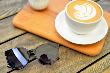 cup of coffee on a wooden stand is on the table in a cafe summer morning, sunglasses in the foreground