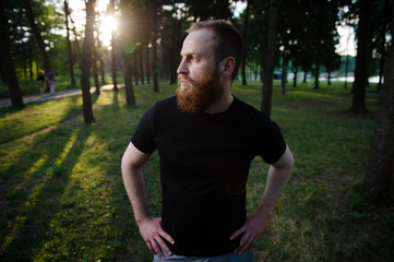 young beautiful bearded caucasian male in black t shirt standing in summer green park during warm sunset with sun ray and lens flare