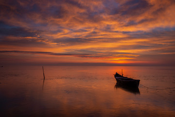 Naklejka premium Colorful sunrise at sea, with a boat floating on the clam water and clouds reflecting on the water