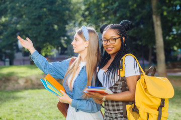 Two happy multiethnic girls as friends with backpaks talking with each other in cheerful way. Cheerful females in university campus park outdoor. Youth Friendship Together Smiling Happiness Concept.