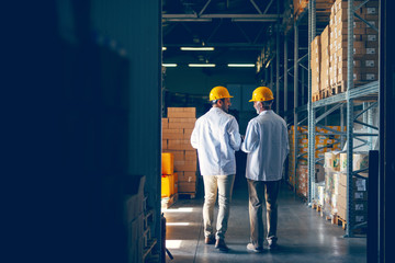 Two smiling warehouse workers in white uniforms and yellow helmets on heads standing and talking about job. Backs turned. - obrazy, fototapety, plakaty