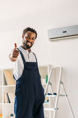 happy african american repairman standing on ladder under air conditioner, looking at camera and showing thumb up