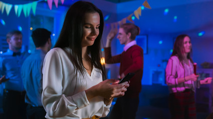 At the Wild House Party: Beautiful Young Girl Uses Smartphone, Browses Through Internet and Social Network. In the Background Crowd of Young People Dancing Off and Having Fun.