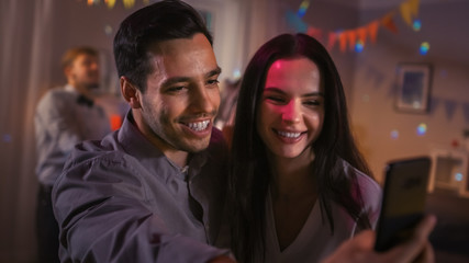 At the Wild House Party: Beautiful Young Couple Take Selfies with a Smartphone for Social Network Sharing. In the Background Crowd of Young People Dancing Off and Having Fun.