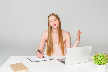 beautiful girl sitting at computer desk, writing in notebook and suffering from heat on grey