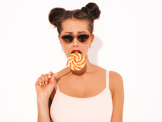 Young beautiful sexy hipster woman with red lips in sunglasses.Trendy girl in summer swimwear clothes. Positive female going crazy.Funny model isolated on white.Two horns.Eating,biting candy lollipop