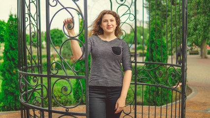 Smiling woman against fence in garden. Content woman looking dreamily standing in green park against ornamental iron fence