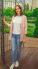 Smiling woman against fence in garden. Content woman looking dreamily standing in green park against ornamental iron fence