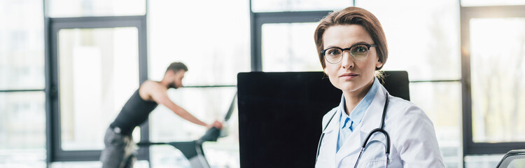 panoramic shot of beautiful doctor in glasses looking at camera near computer in gym