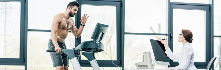panoramic shot of sportsman gesturing on treadmill near doctor during endurance test in gym