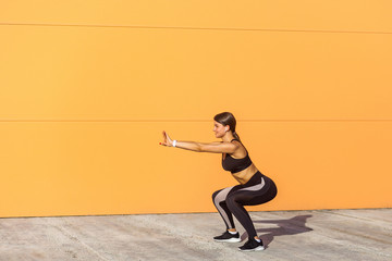 Young attractive woman practicing fitness, doing bodyweight squat exercise, yoga chair pose, working out, wearing black sportswear black pants and top, outdoor full length, on street orange background