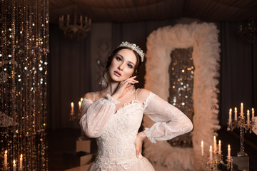 Fototapeta premium Portrait of the bride in the crown. With candles and a mirror. Lace dress. Large feather earrings. Hand on face.