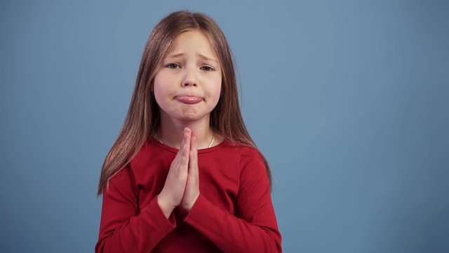 Young beautiful caucasian blonde girl begging saying please praying on blue background in slowmotion. Wearing red sweater. Slow motion