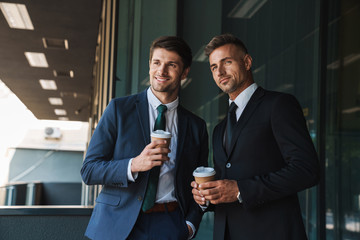 Image of successful businessmen partners talking and drinking takeaway coffee while standing outside office center