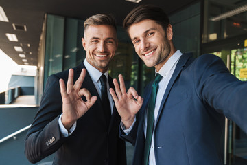 Photo of beautiful two businessmen showing ok sign and taking selfie while standing outside job...