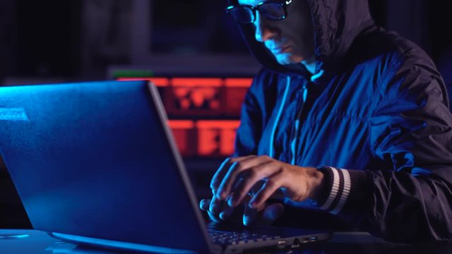 Hacker typing program code while committing a cybercrime hacking a system firewall in the dark under neon light
