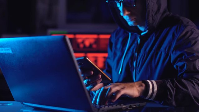 Hacker in the hood holding the phone in his hands trying to hack the mobile device cloud in neon light. Cyber security