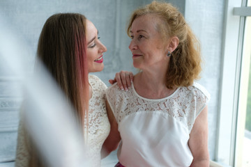 Beautiful old woman with her adult daughter. The woman has blond curly hair, kind eyes and smile. Daughter with long hair and bright lips. Beautiful aging, love, motherhood