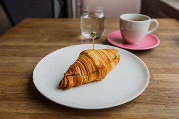 A photo of a croissant with filling on a white plate, a white cup of black coffee on a pink saucer, a glass of water, shot on a wide angle lense.