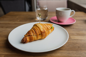 A photo of a croissant with filling on a white plate, a white cup of black coffee on a pink saucer, a glass of water, shot on a wide angle lense.