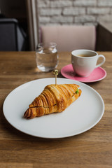 A photo of a croissant with filling on a white plate, a white cup of black coffee on a pink saucer, a glass of water, shot on a wide angle lense.
