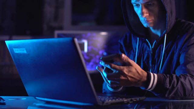 Hacker in the hood holding the phone in his hands trying to hack the mobile device cloud in neon light. Cyber security
