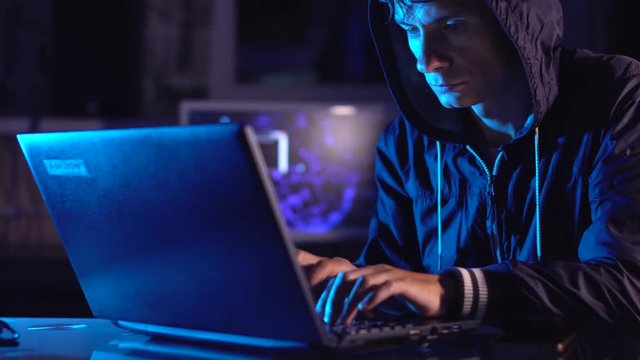 Hacker typing program code while committing a cybercrime hacking a system firewall in the dark under neon light