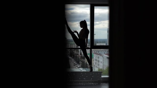Beautiful scene of stretching sporty young woman - girl doing stretch of her legs, raising one foot above her head against the background of large window in slow motion. Silhouette of girl