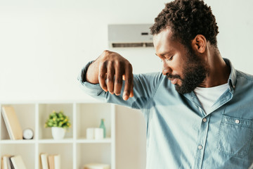 displeased african american man looking at sweaty shirt while suffering from heat at home