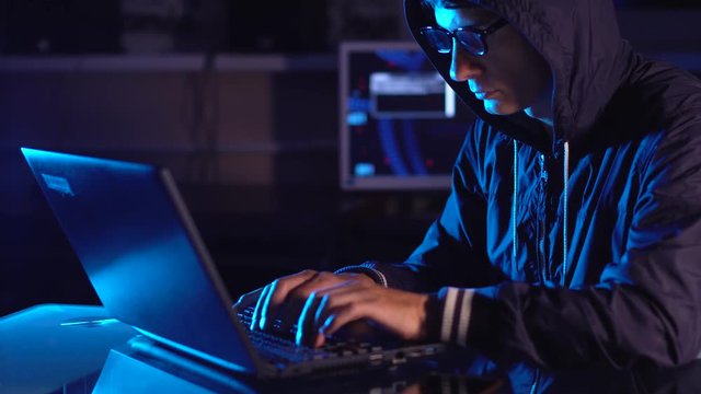 Hacker typing program code while committing a cybercrime hacking a system firewall in the dark under neon light