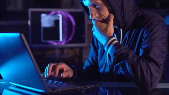 Hacker typing program code while committing a cybercrime hacking a system firewall in the dark under neon light