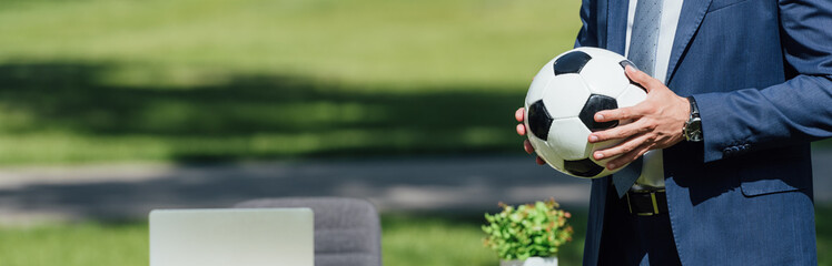 panoramic shot of businessman holding soccer ball in park near table with laptop