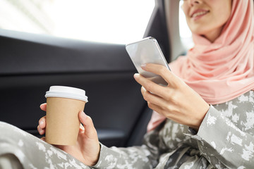Close-up of smiling Muslim woman in hijab sitting in cab and drinking coffee while surfing net on gadget