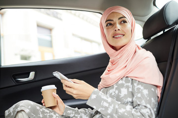 Cheerful attractive young Muslim businesswoman in beautiful headscarf sitting on backseat and using mobile app for taxi ride