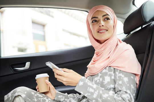Cheerful attractive young Muslim businesswoman in beautiful headscarf sitting on backseat and using mobile app for taxi ride - Powered by Adobe