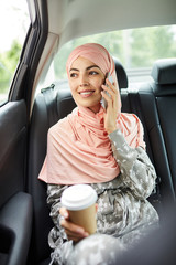 Smiling confident young Muslim woman in pink silk headscarf sitting on backseat of taxi and holding coffee cup while talking on mobile phone