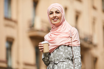 Happy young middle-eastern businesswoman in pink headscarf and summer coat drinking coffee outdoors