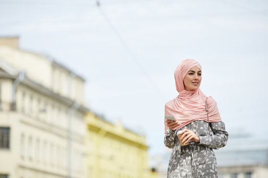 Positive modern young Muslim woman in hijab walking over city street and drinking coffee while using smartphone - Powered by Adobe