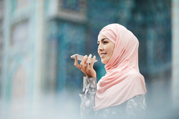 Cheerful confident young middle-eastern woman in pink hijab standing against ornamental building and chatting via voice messages on phone