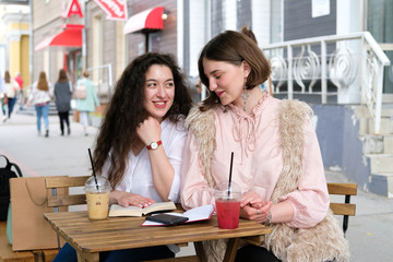 Two young girls sitting at a table in a street cafe. Girlfriends relax and communicate. On the table two summer cocktails in plastic glasses.