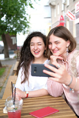Two young girls take a selfie at a table in a street cafe. Girlfriends communicate and have fun. On the table two summer cocktails in plastic glasses.