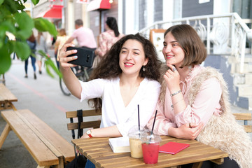 Two young girls take a selfie at a table in a street cafe. Girlfriends communicate and have fun. On the table two summer cocktails in plastic glasses.
