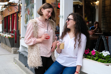 Two young girls are on the street near the summer cafe. Girlfriends communicate and have fun. In the hands of cocktails in plastic glasses.