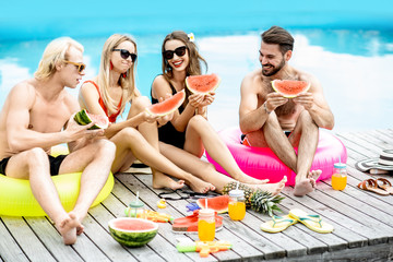 Group of a happy friends in swimwear eating watermelon while sitting together and having fun during the summertime on the swimming pool