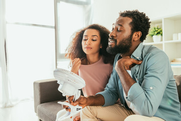 young african man and woman suffering from summer heat and holding blowing electric fan