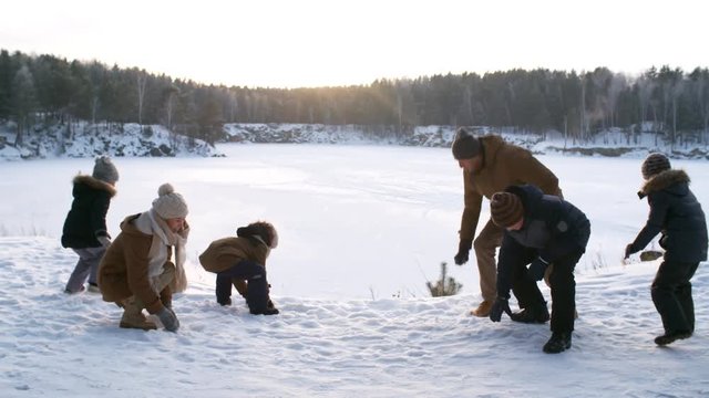 Full shot of active young family with four children having playful snowball fight during scenic winter hike through snowy countryside, tossing up sparkling snow in dim afternoon sunlight