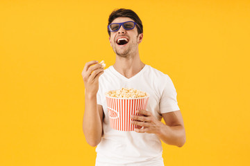 Young man in casual white t-shirt eat popcorn wearing 3d glasses watch film isolated over yellow...